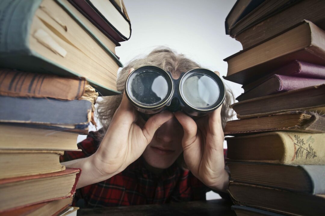 Lady using binoculars while hiding behind large stack of books